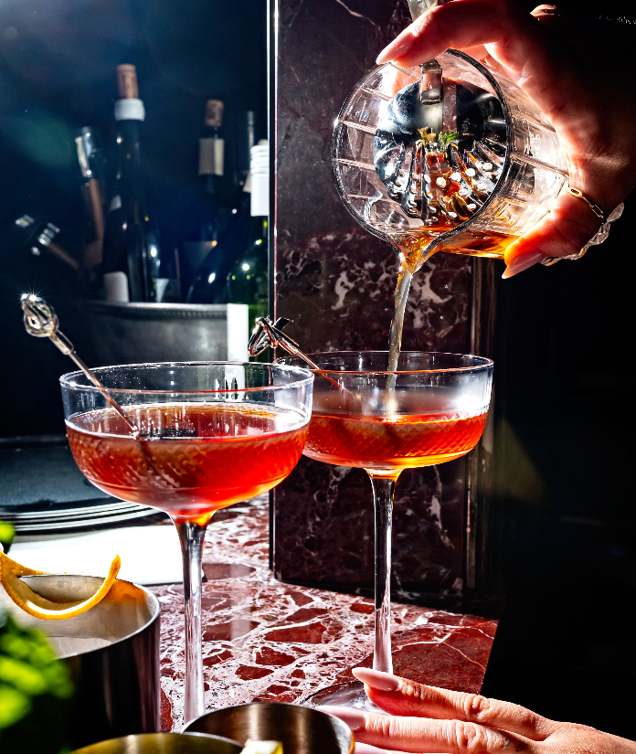 A bartender pouring a red cocktail from a crystal shaker into coupe glasses on a marble bar top.