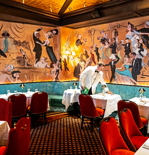 A server setting white-clothed tables in a classic restaurant dining room with red velvet chairs and a vibrant illustrated mural of dancing figures covering the walls.