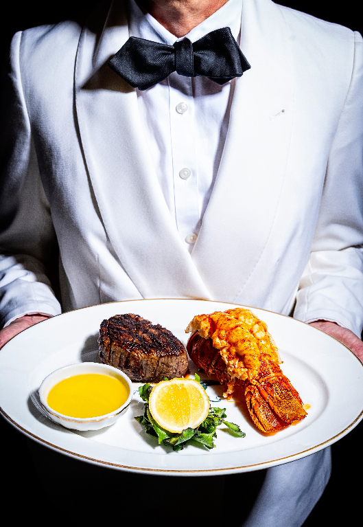 Waiter in white tuxedo jacket holding plate with steak and lobster tail, served with lemon and butter sauce
