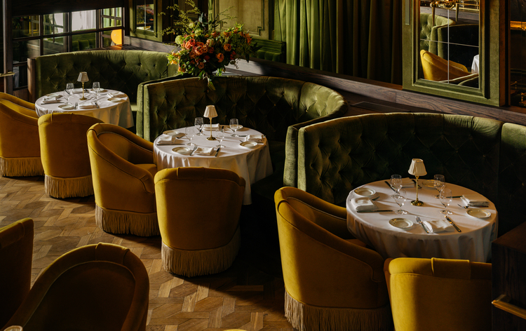 Elegant restaurant interior with green tufted banquettes, yellow armchairs, and round tables set with white tablecloths in a warm, inviting ambiance.